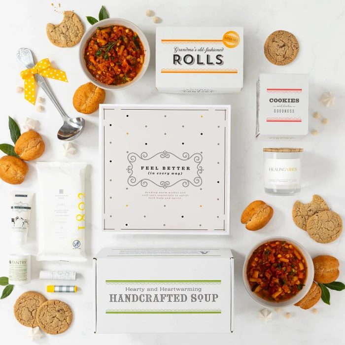 Overhead view of a get-well-themed gift arrangement with a bowl of soup, rolls, cookies, a box labeled “Handcrafted Soup,” a box of rolls, self-care items including a candle and lotion, a metal ladle with a yellow ribbon, and a card with a get-well message.