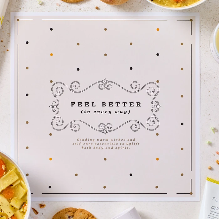 Close-up of a card that reads “Feel Better (in every way),” with decorative accents and parts of a soup and self-care gift arrangement visible around it.