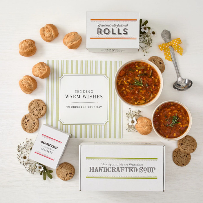 Overhead view of a soup gift arrangement with two bowls of soup, rolls, cookies, a box labeled “Handcrafted Soup,” a box of rolls, a box of cookies, a metal ladle with a yellow ribbon, and a card that reads “Sending Warm Wishes.”
