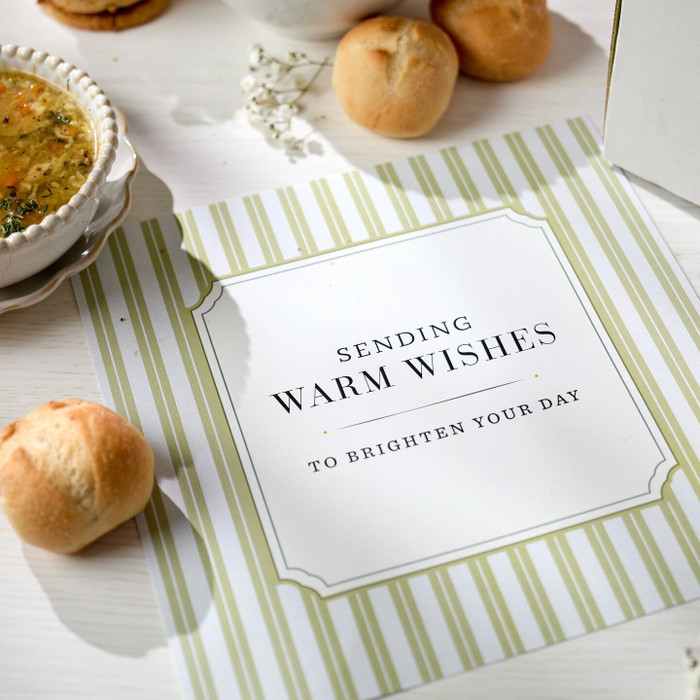 Close-up of a card that reads “Sending Warm Wishes to Brighten Your Day,” placed among rolls and a bowl of soup.