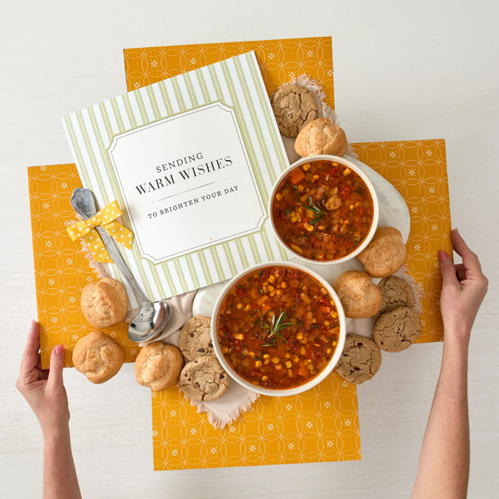 Overhead view of hands holding a yellow gift box arrangement with two bowls of soup, rolls, cookies, a metal ladle with a yellow ribbon, and a card that reads “Sending Warm Wishes.”