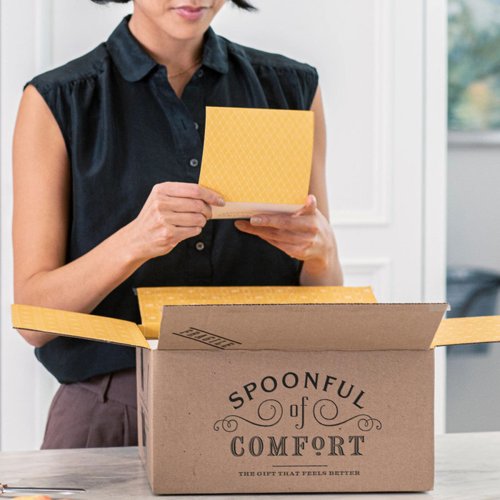 Person opening a Spoonful of Comfort box and holding a yellow card, standing at a table indoors.