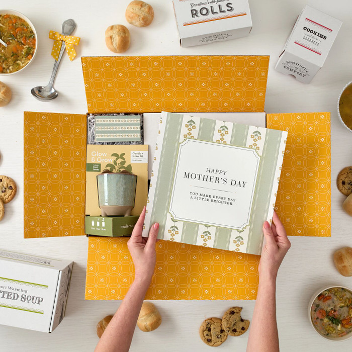 Overhead view of hands holding a Mother’s Day gift arrangement with a candle grow kit, a card that reads “Happy Mother’s Day,” and surrounding items including soup, rolls, cookies, and packaged goods on a yellow background.