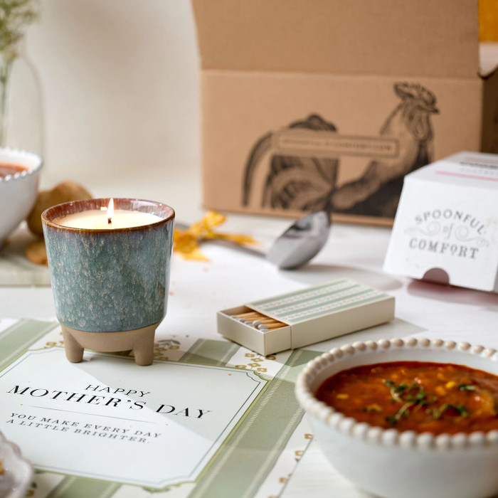 Close-up of a lit candle on a Mother’s Day card, with a matchbox, a bowl of soup, and a ladle in the background.