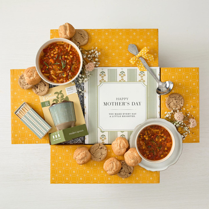 Overhead view of a yellow gift box arrangement with two bowls of soup, rolls, cookies, a candle gift set, a metal ladle with a yellow ribbon, and a card that reads “Happy Mother’s Day.”