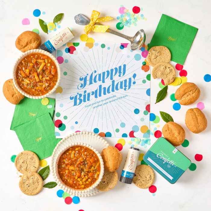 Overhead view of a birthday-themed arrangement with two bowls of soup, rolls, cookies, colorful confetti, green napkins, confetti poppers, and a card that reads “Happy Birthday!”