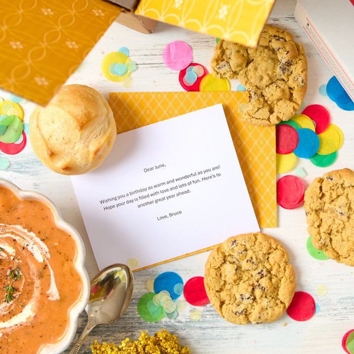 Close-up of a birthday card placed among a bowl of soup, rolls, cookies, and colorful confetti on a yellow background.