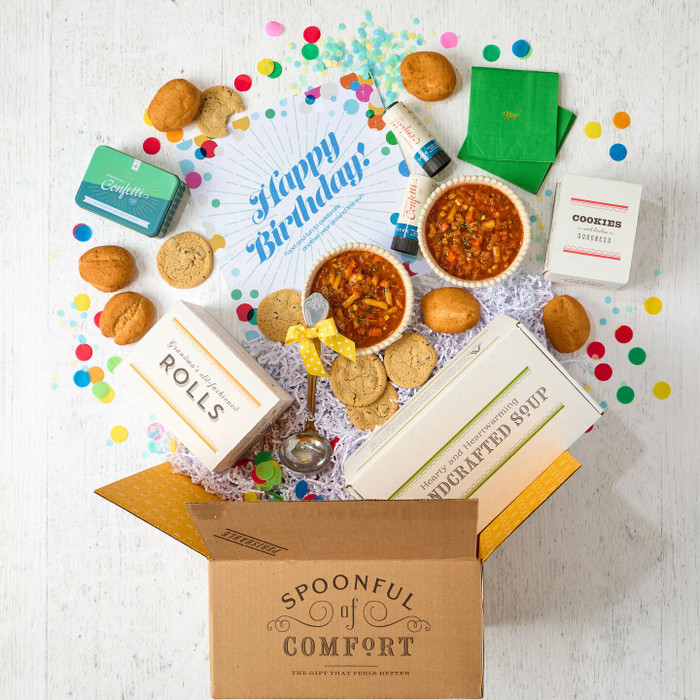 Open Spoonful of Comfort gift box with a birthday-themed arrangement including two bowls of soup, rolls, cookies, colorful confetti, green napkins, confetti poppers, packaged items, and a card that reads “Happy Birthday!”