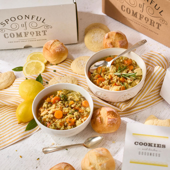 Overhead view of two bowls of soup with rolls, cookies, lemons, and a spoon on a yellow striped cloth, with Spoonful of Comfort boxes in the background.