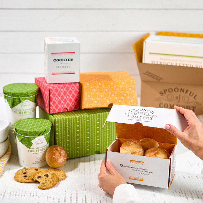 Hands holding an open gift box filled with soup, bread, cookies, and packaged comfort food products.