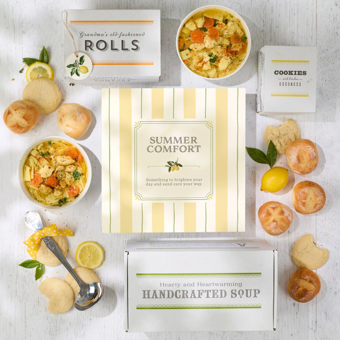 Overhead view of a comfort food gift box with soup mixes, rolls, cookies, and prepared soup bowls displayed on a marble patterned background.