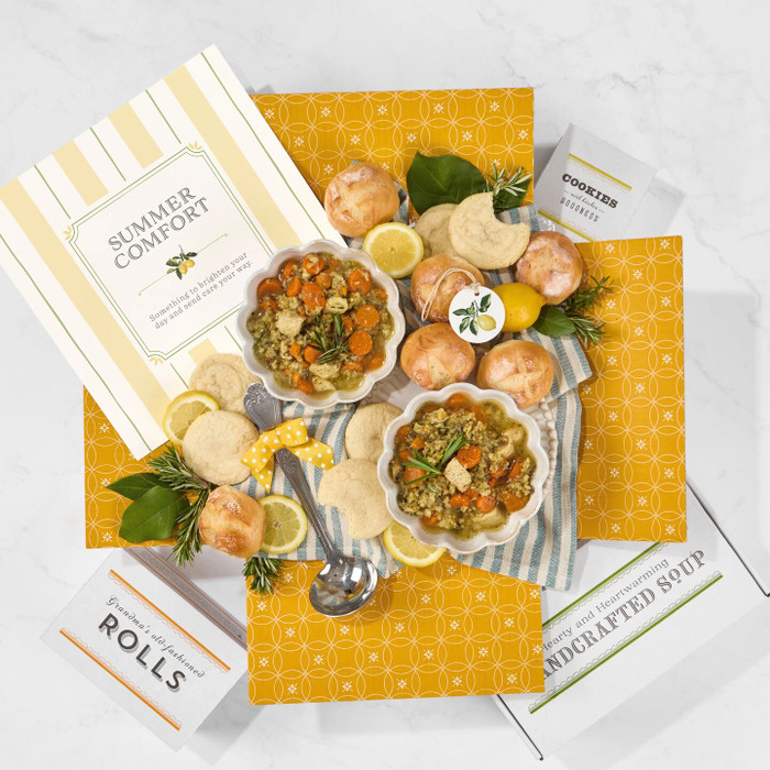 Overhead view of a comfort food gift box featuring packaged soup mixes, rolls, cookies, and prepared soup bowls arranged on a yellow patterned background. A “Summer Comfort” card is positioned in the box