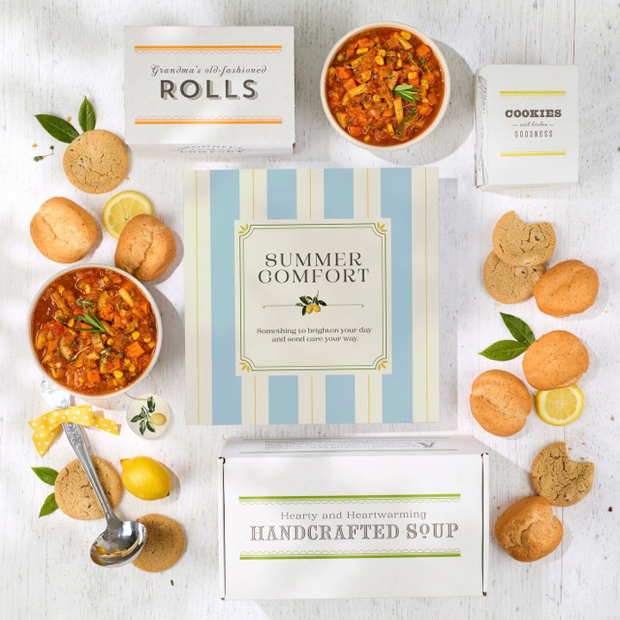 Overhead view of a summer-themed gift arrangement with two bowls of soup, rolls, cookies, lemons, a container labeled “Handcrafted Soup,” a metal ladle with a yellow ribbon, and a card that reads “Summer Comfort.”