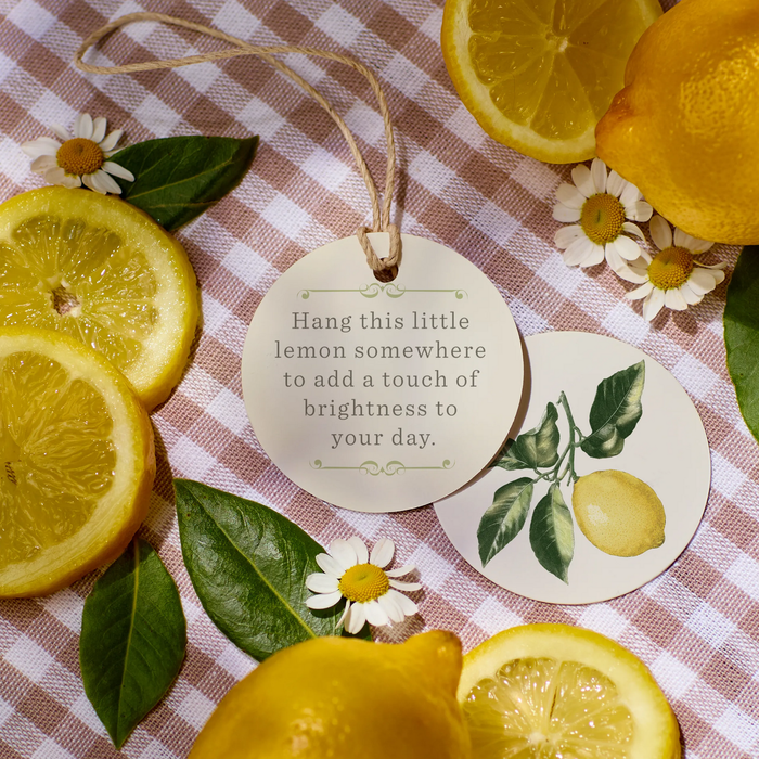 Lemon-themed gift tag that reads “Hang this little lemon somewhere to add a touch of brightness to your day,” surrounded by fresh lemons, slices, greenery, and small white flowers on a checkered cloth.