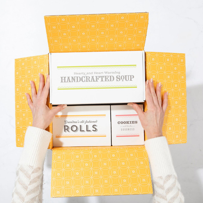 hands opening a box, with a soup box, rolls box, and cookie box showing in the open box