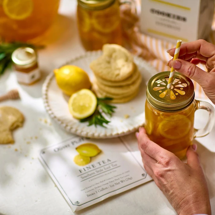 Close-up of hands placing a striped paper straw into a glass mason jar of iced tea with lemon slices, with cookies, a lemon, honey, and a package of tea in the background.
