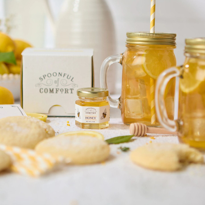 Gift set arranged on a table featuring an iced tea sachet, lemon cookies in a yellow box, two glass mason jar mugs, yellow-and-white striped paper straws, a mini honey jar with a wooden dipper, a yellow-striped kitchen towel, a lemon gift tag, and a card that reads “Sweet Summertime.”