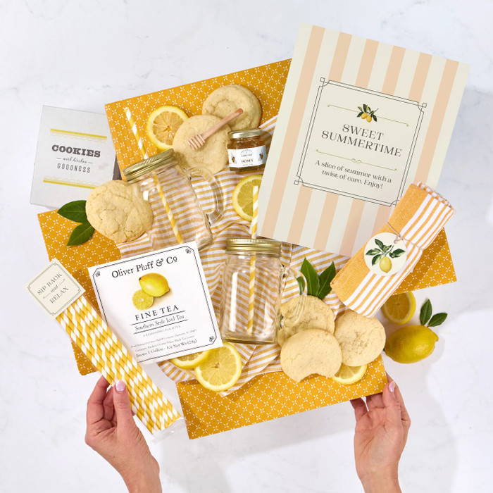 Overhead view of hands holding a summer-themed gift arrangement with lemon cookies, two glass mason jars, lemon slices, a jar of honey, striped paper straws, a kitchen towel, and a card that reads “Sweet Summertime.”