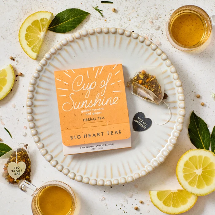Close-up of a package of “Cup of Sunshine” herbal tea placed on a plate, surrounded by lemon slices, tea, a honey dipper, and small decorative items.