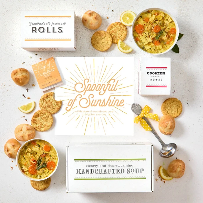 Overhead view of a sunshine-themed gift arrangement with two bowls of soup, rolls, cookies, lemon slices, a box labeled “Handcrafted Soup,” a box of rolls, a box of cookies, a metal ladle with a yellow ribbon, and a card that reads “Spoonful of Sunshine.”