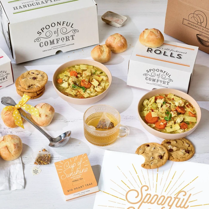 Soup gift set displayed with two bowls of soup, rolls, cookies, Spoonful of Comfort boxes, a glass of tea, a metal ladle with a yellow ribbon, and a card that reads “Spoonful of Sunshine.”
