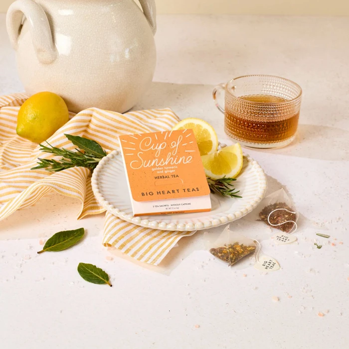 Package of “Cup of Sunshine” herbal tea placed on a plate with lemon slices, next to a glass of tea, a teapot, and a yellow striped towel.