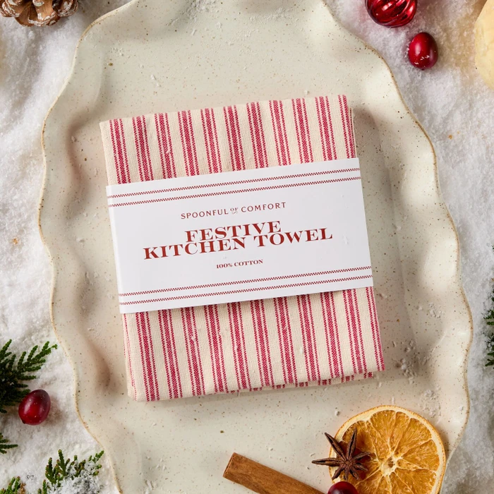 Folded red and cream striped Spoonful of Comfort Festive Kitchen Towel, made from 100% cotton, wrapped in a branded paper band and styled on a rustic cream platter with festive greenery, cranberries, dried orange, and cinnamon.