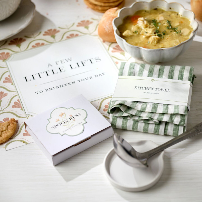 Close-up of a spring-themed gift arrangement featuring a card that reads “A Few Little Lifts to Brighten Your Day,” a bowl of soup, a green striped kitchen towel labeled “At Your Service,” and a metal ladle resting on a stand.