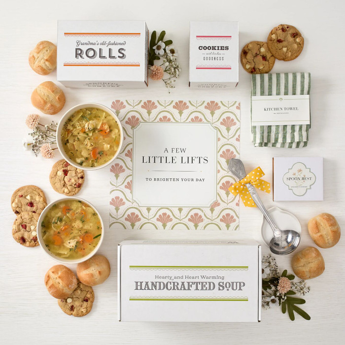 Overhead view of a spring-themed gift arrangement with two bowls of soup, rolls, cookies, a box labeled “Handcrafted Soup,” a box of rolls, a box of cookies, a green striped kitchen towel, a metal ladle with a yellow ribbon, greenery, and a card that reads “A Few Little Lifts.”