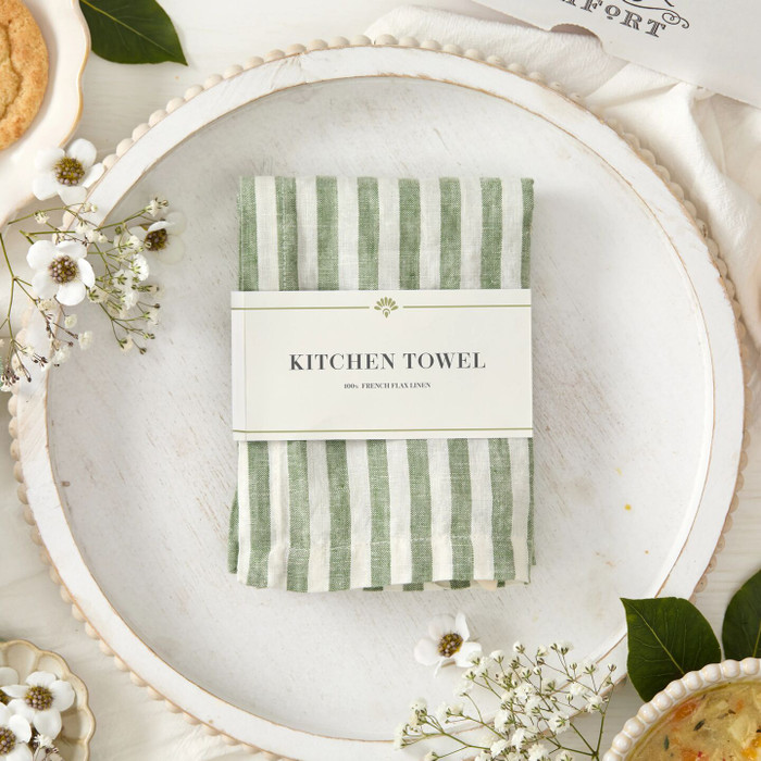 Close-up of a folded green striped kitchen towel labeled “Kitchen Towel,” placed on a plate with small white flowers and greenery nearby.
