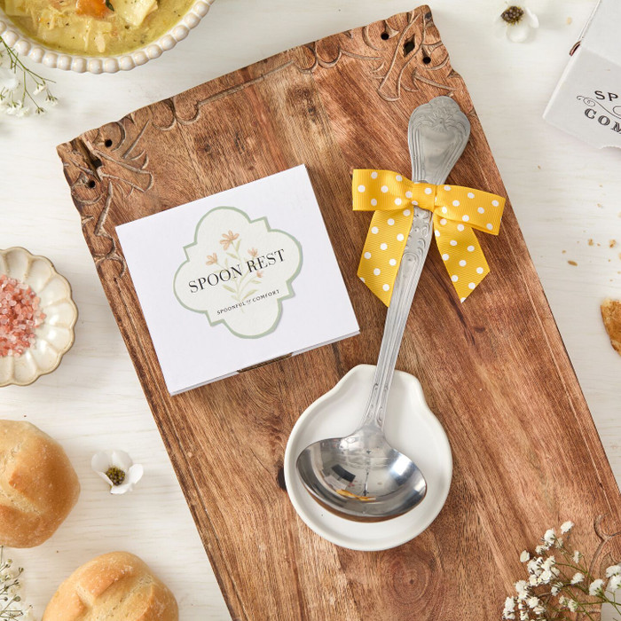 Close-up of a metal ladle tied with a yellow ribbon, resting on a white spoon rest on a wooden board, next to the product box labeled “Spoon Rest,” with rolls and small flowers nearby.