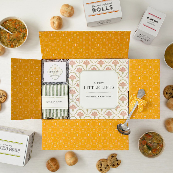 Open yellow gift box displaying a spring-themed arrangement with a card that reads “A Few Little Lifts,” a green striped kitchen towel, a spoon rest box, and surrounding items including soup, rolls, cookies, and packaged goods.