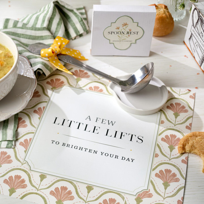Close-up of a spring-themed card that reads “A Few Little Lifts to Brighten Your Day,” surrounded by a bowl of soup, a green striped towel, a spoon, and a metal ladle with a yellow ribbon.