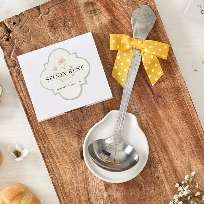 Close-up of a metal ladle tied with a yellow ribbon, resting on a wooden board with a spoon rest, alongside a small “Spoon Rest” card and surrounding rolls and flowers.