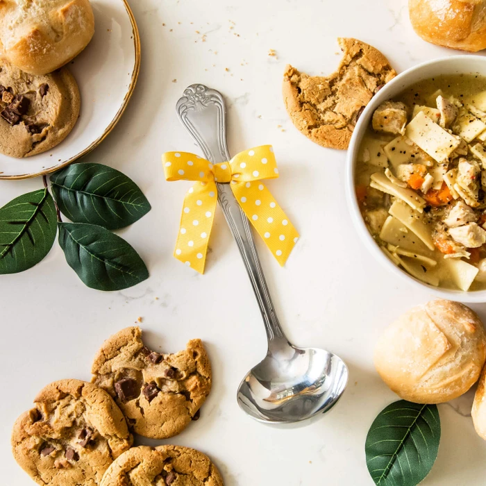 Overhead view of a metal ladle with a yellow ribbon surrounded by bowls of soup, rolls, cookies, and green leaves on a white surface.