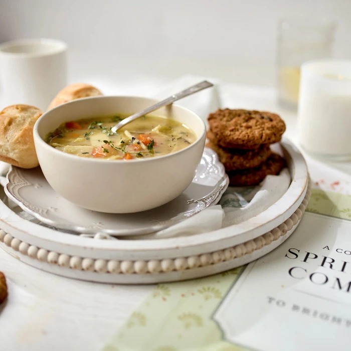 Close-up of a bowl of chicken soup on a tray with a roll, cookies, and a glass of milk, with a spring-themed card partially visible nearby.