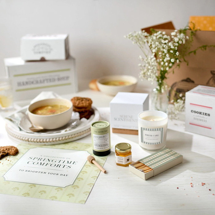 Spring-themed gift arrangement displayed with bowls of soup, rolls, cookies, boxes labeled “Handcrafted Soup” and “Cookies,” jasmine green tea, a mini jar of honey with a dipper, a candle with a box of matches, and a card that reads “Springtime Comfort.”