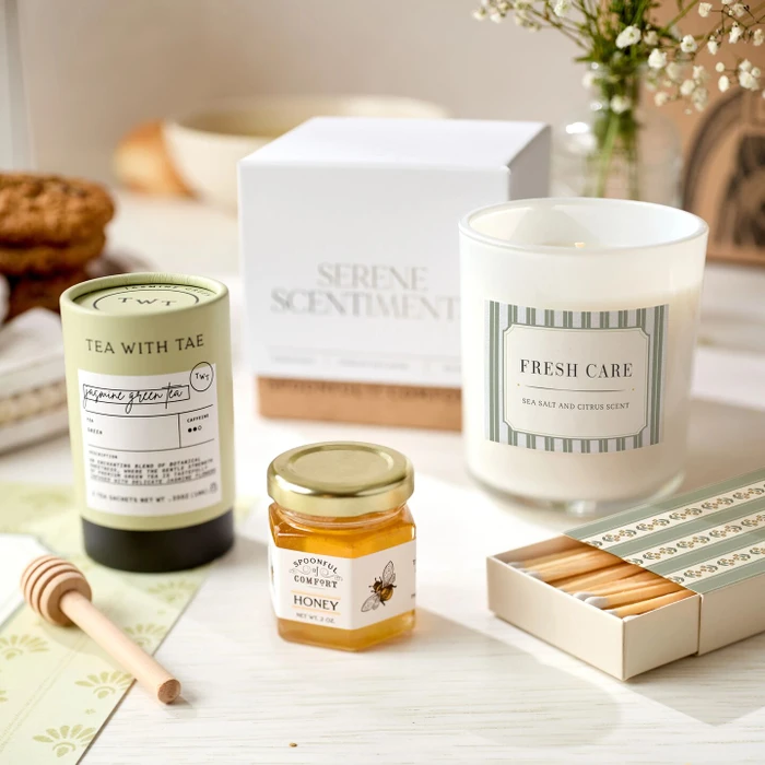 Close-up of self-care items including jasmine green tea, a mini jar of raw honey with a wooden dipper, a “Fresh Care” candle, and a coordinating box of matches, with a “Serene Scents” box in the background.