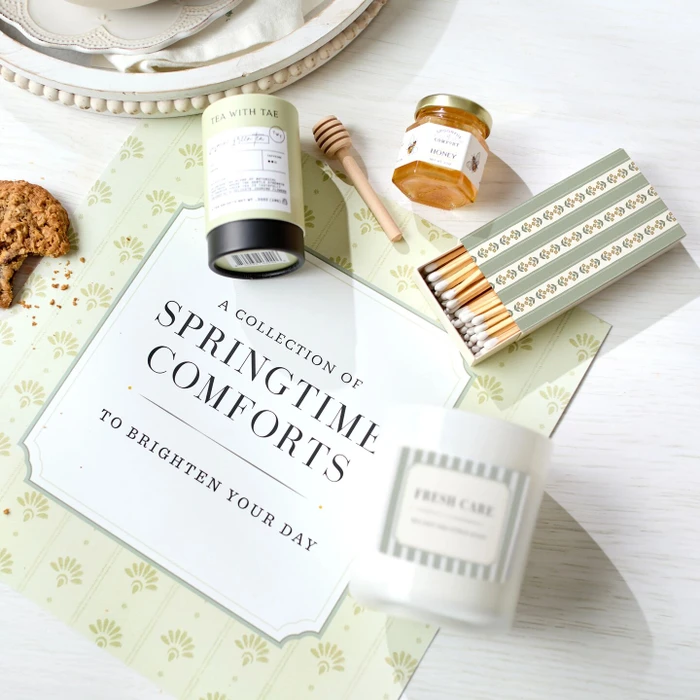 Close-up of a spring-themed card that reads “A Collection of Springtime Comforts to Brighten Your Day,” surrounded by jasmine green tea, a mini jar of honey with a dipper, a candle, and a box of matches.