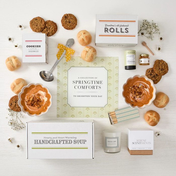 Overhead view of a spring-themed gift arrangement with two bowls of tomato soup, rolls, cookies, a box labeled “Handcrafted Soup,” a box of rolls, a box of cookies, jasmine green tea sachets, raw honey with a mini dipper, a lemon and sea salt candle with a coordinating box of matches, a metal ladle with a yellow ribbon, greenery, and a card that reads “Springtime Comfort.”