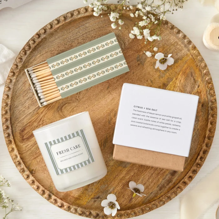Close-up of a candle labeled “Fresh Care” next to its product box and a coordinating box of matches, arranged on a wooden tray with small white flowers.