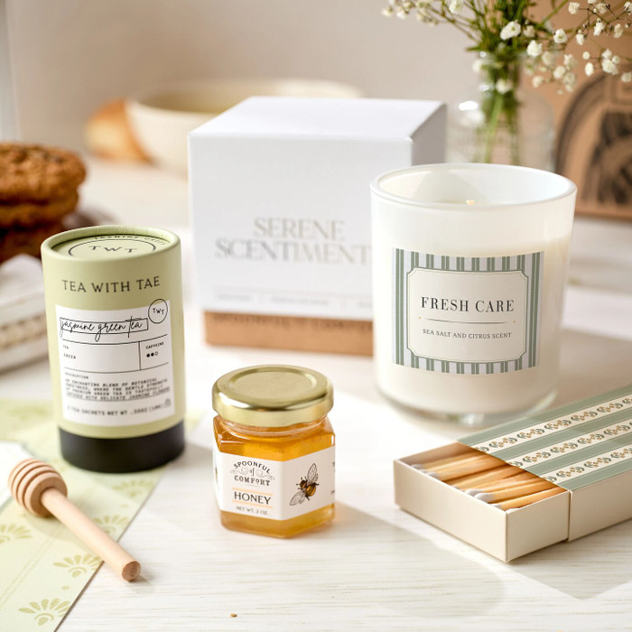Close-up of self-care items including jasmine green tea, a mini jar of raw honey with a wooden dipper, a “Fresh Care” candle, and a coordinating box of matches, with a “Serene Scents” box in the background.