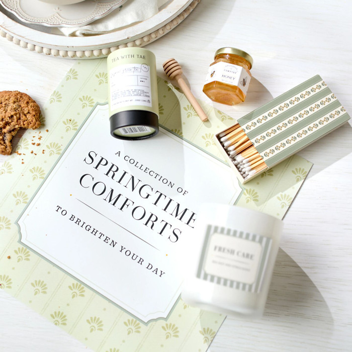 Close-up of a spring-themed card that reads “A Collection of Springtime Comforts to Brighten Your Day,” surrounded by jasmine green tea, a mini jar of honey with a dipper, a candle, and a box of matches.