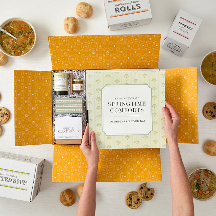 Overhead view of hands holding a spring-themed gift arrangement with a card that reads “Springtime Comforts,” alongside jasmine green tea, a candle with matches, and surrounding bowls of soup, rolls, cookies, and packaged goods.