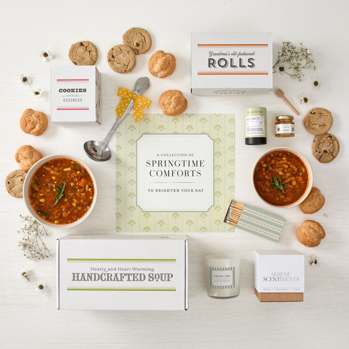 Overhead view of a spring-themed gift arrangement with two bowls of tomato soup, rolls, cookies, a box labeled “Handcrafted Soup,” a box of rolls, a box of cookies, jasmine green tea sachets, raw honey with a mini dipper, a lemon and sea salt candle with a coordinating box of matches, a metal ladle with a yellow ribbon, greenery, and a card that reads “Springtime Comfort.”