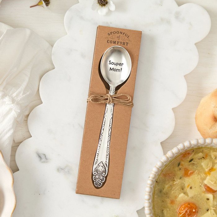Close-up of a packaged spoon engraved “Souper Mom!” tied with twine, placed on a marble surface next to a bowl of soup.