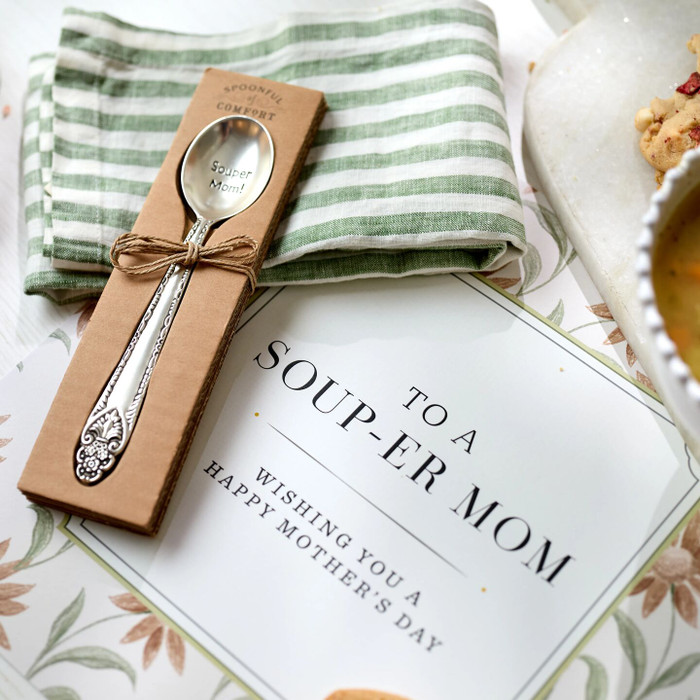 Close-up of a packaged spoon engraved “Souper Mom!” resting on a green striped cloth beside a card that reads “To a Soup-er Mom. Wishing you a Happy Mother’s Day.”
