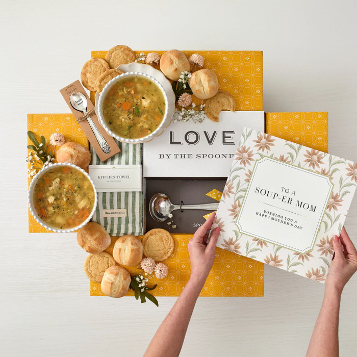 Overhead view of hands holding a Mother’s Day gift arrangement with two bowls of soup, rolls, cookies, a container labeled “Handcrafted Soup,” a metal ladle with a yellow ribbon, a green striped towel, and cards that read “To a Soup-er Mom”