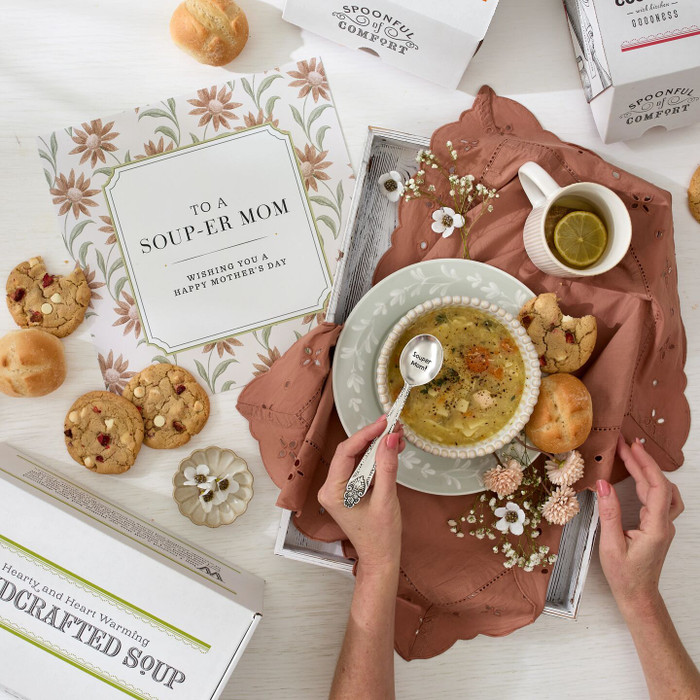 Overhead view of hands holding a bowl of soup on a tray with rolls, cookies, a small cup of tea, and a card that reads “To a Soup-er Mom,” with additional packaged items surrounding the scene.