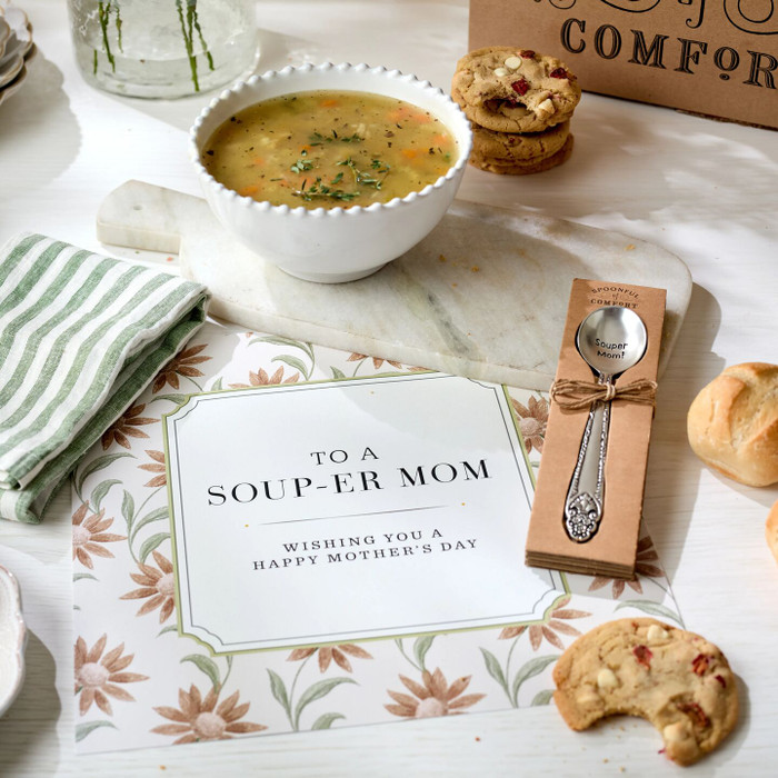 Close-up of a soup gift arrangement with a bowl of soup, cookies, a green striped towel, a packaged “Souper Mom!” spoon, and a card that reads “To a Soup-er Mom. Wishing you a Happy Mother’s Day.”